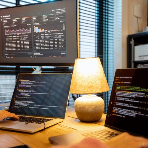 Female programmer writing programming code on laptops and desktop computer at cozy home workplace. Close-up on hands and keyboard